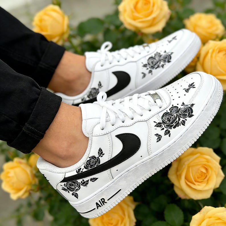May include: White sneakers with black rose floral designs and a black swoosh logo. The word "AIR" is printed on the side of the shoe. The shoes are laced with white laces.