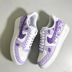 May include: Pair of white and light purple sneakers with a grape-themed design. The sneakers feature a white sole, white laces, and a purple swoosh. The design includes purple grape clusters and dripping purple accents.