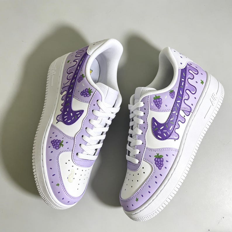 May include: Pair of white and light purple sneakers with a grape-themed design. The sneakers feature a white sole, white laces, and a purple swoosh. The design includes purple grape clusters and dripping purple accents.