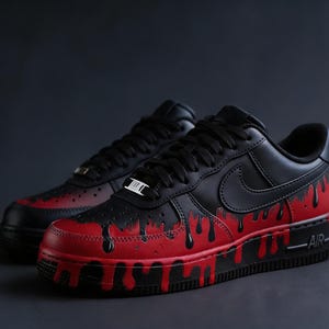 May include: Pair of black and red Air Force 1 sneakers. The shoes have a black upper with a red base and dripping paint design. The Nike swoosh is visible on the side. The laces are black.
