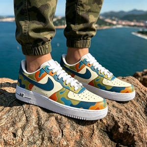 May include: A pair of Air Force 1 sneakers with a cream-colored base and a colorful camouflage pattern in blue, orange, and green. The shoes have a white sole and laces, with a blue Nike swoosh.