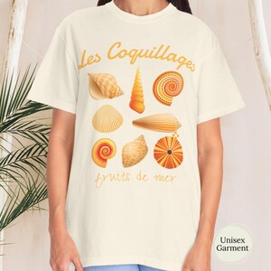 May include: Cream-coloured unisex t-shirt with a design of various seashells in orange and tan hues. The French text "Les Coquillages fruits de mer" is printed above and below the illustrations. The shirt is a unisex garment.