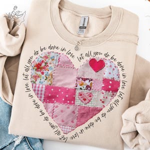 May include: Beige sweatshirt with a patchwork heart design. The heart is composed of various pink fabrics, including floral and polka dot patterns. The phrase "Let all you do be done in love" is printed around the heart. A silver necklace is draped on the sweatshirt.