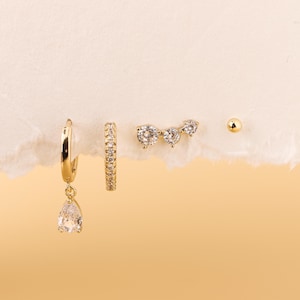 May include: A collection of gold-toned earrings displayed on a textured white surface against a soft yellow background. The set includes a pair of hoop earrings with a teardrop-shaped charm, a pair of small hoop earrings, a triple-stone stud, and a small ball stud.