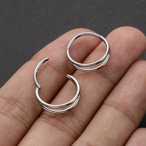 Può includere: Due orecchini a cerchio doppi color argento. Uno è aperto, rivelando la chiusura, l'altro è chiuso. Gli orecchini sono circolari con un secondo cerchio più piccolo all'interno di quello più grande.