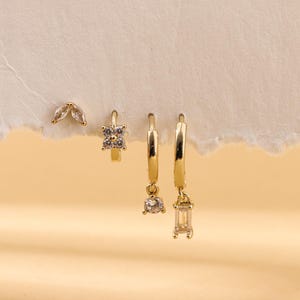 Może przedstawiać: Zestaw złotych kolczyków z błyszczącymi detalami. Zestaw zawiera kolczyki sztyfty z motywami liści, małe kolczyki koła z kwadratowymi kamieniami oraz wiszące kolczyki koła z okrągłymi i prostokątnymi kamieniami. Prezentowane na teksturowanej, kremowej powierzchni.