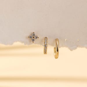 May include: A set of three gold earrings. One is a stud with a four-petal design, another is a small hoop with clear stones, and the third is a plain gold hoop. The earrings are displayed on a textured, off-white surface.