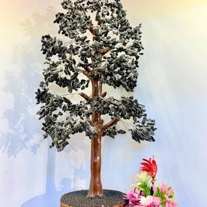 Pode incluir: Uma escultura decorativa de árvore com tronco e galhos marrons, adornada com folhas cinza escuro. A árvore está sobre uma base redonda e texturizada. Um pequeno arranjo de flores artificiais coloridas em um vaso branco é colocado próximo. Decoração de casa única.