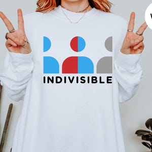 May include: White sweatshirt with the word "INDIVISIBLE" in black font. The graphic above the text features blue, red, and gray abstract figures. The sweatshirt has long sleeves and a crew neck.