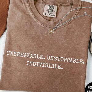 May include: Espresso-colored t-shirt with the words "UNBREAKABLE. UNSTOPPABLE. INDIVISIBLE." printed in white. The shirt has a crew neck and short sleeves. A silver necklace with a pendant is draped across the shirt.
