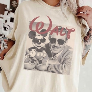 May include: An ivory-colored crew neck t-shirt featuring a graphic print of Mickey Mouse and Walt Disney both wearing sunglasses. The word "WALT" is printed in large, faded red letters above the figures. The shirt has short sleeves and a relaxed fit.