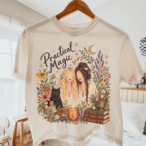 May include: An ivory short-sleeve t-shirt featuring a graphic print with the text "Practical Magic" above two women, a black cat, a candle, a mortar and pestle, and a stack of books, all surrounded by colorful floral and botanical illustrations.