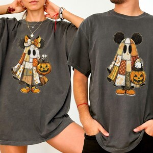 May include: A charcoal gray short-sleeve t-shirt featuring a graphic of a ghost wearing a patchwork-style blanket with a pumpkin-shaped trick-or-treat bucket. The ghost has mouse-like ears and is holding the orange pumpkin bucket in one hand.