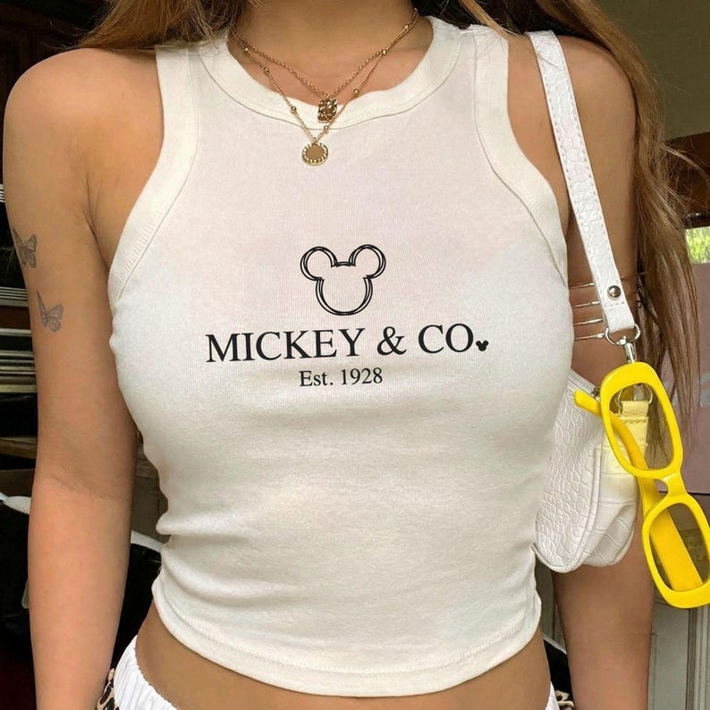 以下が含まれることがあります： 黒色の「MICKEY & CO. Est. 1928」の文字が入った白いリブタンクトップ。ミッキーマウスの頭のグラフィックが特徴です。白いストラップの白いハンドバッグと黄色のサングラスも表示されています。