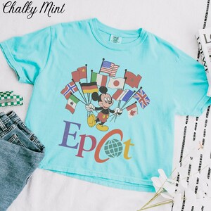 Op de afbeelding: Een lichtblauw T-shirt met Mickey Mouse die vlaggen uit de hele wereld vasthoudt. Het woord "Epcot" staat eronder gedrukt. Het shirt wordt getoond met een spijkerbroek en een klein groen-wit geruit item.