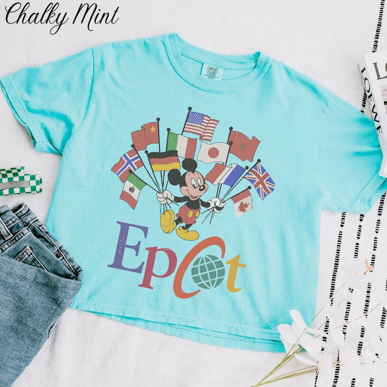 Op de afbeelding: Een lichtblauw T-shirt met Mickey Mouse die vlaggen uit de hele wereld vasthoudt. Het woord "Epcot" staat eronder gedrukt. Het shirt wordt getoond met een spijkerbroek en een klein groen-wit geruit item.