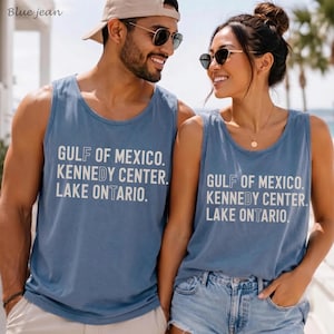 May include: Two matching blue tank tops featuring white text that reads GULF OF MEXICO, KENNEDY CENTER, LAKE ONTARIO. The shirts have a crew neckline and a sleeveless design. The text is printed in a clean, sans-serif font across the chest area.