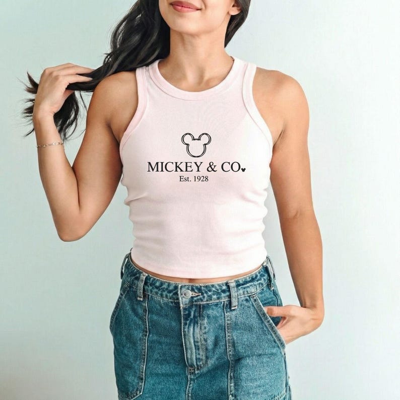 以下が含まれることがあります： 「MICKEY & CO.」と「Est. 1928」の文字が黒でプリントされた、薄いピンク色のリブ編みタンクトップ。タンクトップはノースリーブで、丸いネックラインです。モデルは青いデニムジーンズを着用しています。