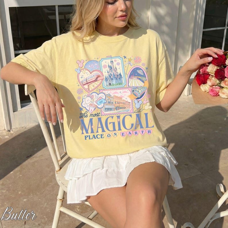 Pu&ograve; includere: T-shirt giallo chiaro con un design grafico colorato raffigurante un castello e un treno. Il testo "The most MAGICAL PLACE ON EARTH" &egrave; stampato. La persona indossa una gonna bianca.