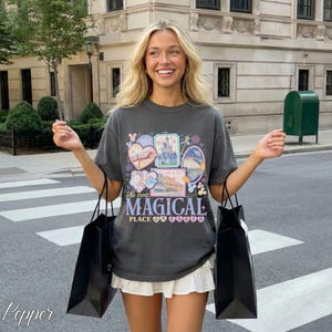 Pu&ograve; includere: T-shirt grigio scuro con una grafica colorata con cuori, castelli e la scritta "The Most Magical Place". La maglietta &egrave; abbinata a una gonna bianca e borse della spesa nere. L'immagine &egrave; scattata all'aperto.