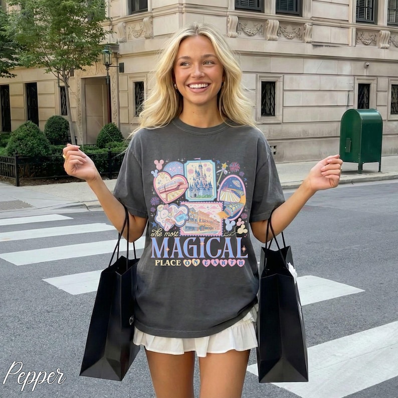 Pu&ograve; includere: T-shirt grigio scuro con una grafica colorata con cuori, castelli e la scritta "The Most Magical Place". La maglietta &egrave; abbinata a una gonna bianca e borse della spesa nere. L'immagine &egrave; scattata all'aperto.