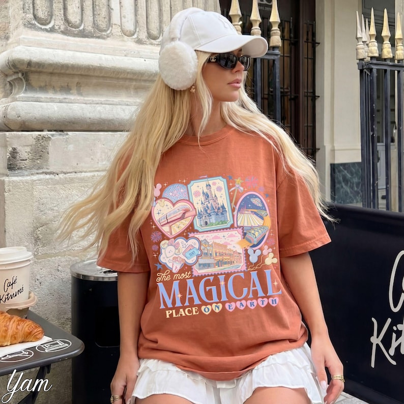 Pu&ograve; includere: T-shirt color ruggine con un design grafico colorato con varie immagini e la scritta "The most MAGICAL PLACE ON EARTH". Il design include un castello, un cuore e altri elementi a tema. La maglietta &egrave; a maniche corte.