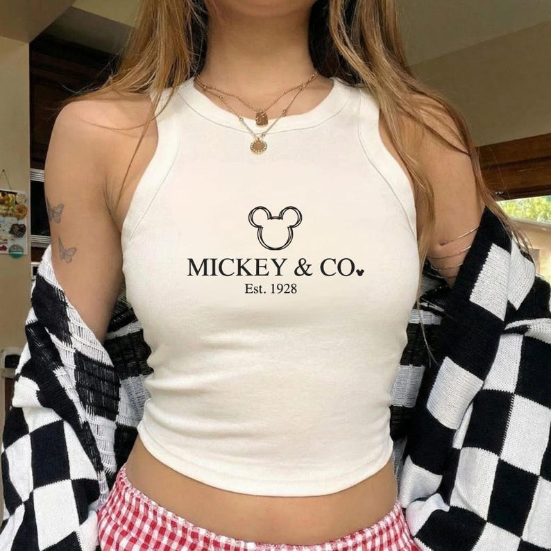 以下が含まれることがあります： 「MICKEY & CO.」の文字と「Est. 1928」の年号が黒でプリントされたクリーム色のタンクトップ。 ミッキーマウスの頭のグラフィックが特徴です。 黒と白のチェック柄のジャケットが肩にかけられています。