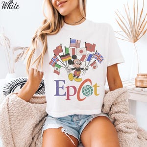Op de afbeelding: Wit t-shirt met Mickey Mouse die vlaggen van verschillende landen vasthoudt. Het woord "Epcot" is in kleurrijke letters onder de afbeelding gedrukt. Het woord "White" staat in de linkerbovenhoek.