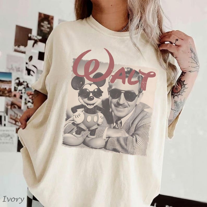 May include: An ivory-colored crew neck t-shirt featuring a graphic print of Mickey Mouse and Walt Disney both wearing sunglasses. The word "WALT" is printed in large, faded red letters above the figures. The shirt has short sleeves and a relaxed fit.