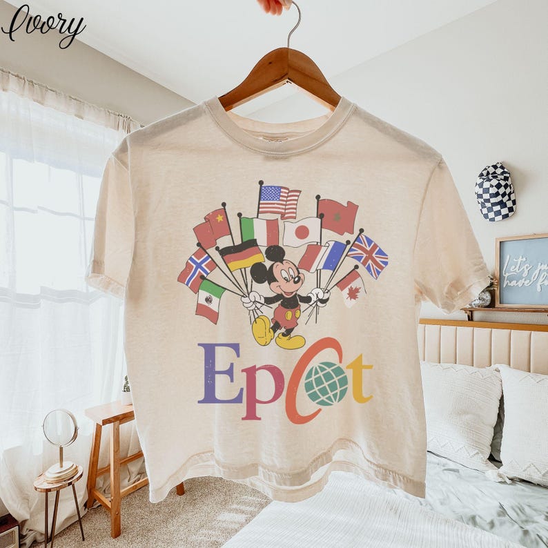 Op de afbeelding: Cr&egrave;mekleurig T-shirt met Mickey Mouse die vlaggen van verschillende landen vasthoudt. Het woord "Epcot" is in kleurrijke letters onder de afbeelding gedrukt. Het shirt hangt aan een houten hanger.