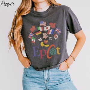 Op de afbeelding: Donkergrijs t-shirt met Mickey Mouse die vlaggen van over de hele wereld vasthoudt. Het woord "Epcot" is in kleurrijke letters onder de afbeelding gedrukt. Het shirt heeft een vintage stijl.