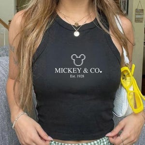 以下が含まれることがあります： 「MICKEY & CO.」と「Est. 1928」の文字が白でプリントされた黒のタンクトップ。丸いネックラインとノースリーブのデザインです。重ね着のネックレスと白いハンドバッグが合わせられています。