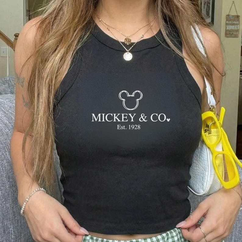 以下が含まれることがあります： 「MICKEY & CO.」と「Est. 1928」の文字が白でプリントされた黒のタンクトップ。丸いネックラインとノースリーブのデザインです。重ね着のネックレスと白いハンドバッグが合わせられています。