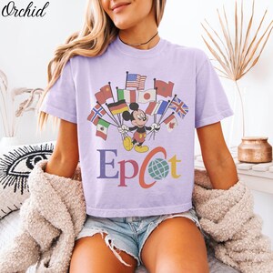 Op de afbeelding: Lichtpaars T-shirt met Mickey Mouse die vlaggen van verschillende landen vasthoudt. Het woord "Epcot" staat eronder in verschillende kleuren. Het shirt heeft korte mouwen en een ronde hals.
