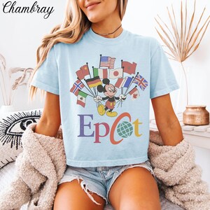 Op de afbeelding: Lichtblauw T-shirt met Mickey Mouse die vlaggen van verschillende landen vasthoudt. Het woord "Epcot" staat eronder in verschillende kleuren gedrukt. Het T-shirt heeft korte mouwen en een losse pasvorm.