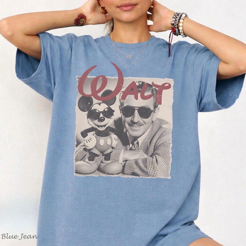 May include: A blue short-sleeve t-shirt featuring a graphic print of Walt Disney and Mickey Mouse wearing sunglasses. The text "Walt" is printed in a large, stylized maroon font above the black and white image of the two figures.