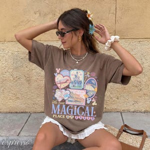 Pu&ograve; includere: T-shirt marrone con una grafica colorata con la scritta "The most MAGICAL PLACE ON EARTH". Il design include immagini di un treno, un castello e altri elementi del parco a tema. La maglietta &egrave; indossata da una persona.