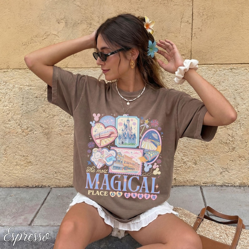 Pu&ograve; includere: T-shirt marrone con una grafica colorata con la scritta "The most MAGICAL PLACE ON EARTH". Il design include immagini di un treno, un castello e altri elementi del parco a tema. La maglietta &egrave; indossata da una persona.