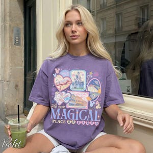 Pu&ograve; includere: Una maglietta viola con un design grafico che presenta varie illustrazioni, tra cui un castello, un treno e altri elementi a tema. Il testo "The most MAGICAL PLACE ON EARTH" &egrave; visualizzato sul davanti. La parola "Violet" &egrave; scritta in basso a sinistra.