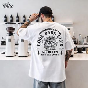 May include: White t-shirt with a graphic on the back that reads "COOL DADS CLUB" with a cartoon coffee cup wearing sunglasses. The text "NO RULES JUST DAD JOKES" is also printed on the shirt. The word "White" is printed on the top left.