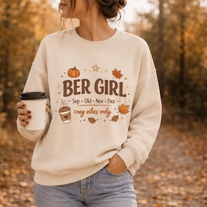 Könnte beinhalten: Ein cremefarbenes Sweatshirt mit Rundhalsausschnitt, braunem Schriftzug BER GIRL, den Monaten Sep, Okt, Nov, Dez und dem Text cozy vibes only. Das Design zeigt Illustrationen eines Kürbisses, Herbstblättern, Sternen und einem Kaffeebecher.