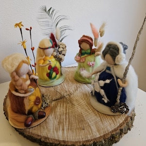 May include: Four whimsical felted figures, each with unique attire and accessories, are arranged on a rustic wooden slice. The figures showcase a variety of colors, including yellow, green, blue, and brown, and are adorned with decorative elements like flowers and twigs.