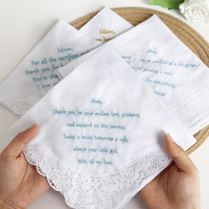 Pode incluir: Lenços brancos com mensagens bordadas em verde-azulado e dourado. Um lenço é segurado, exibindo uma mensagem para "Mãe" com uma borda de renda. Outros lenços têm mensagens para "Mãe" e "Pai".