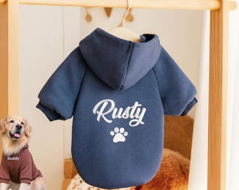 Może przedstawiać: Granatowa bluza z kapturem dla psa z kapturem, wisząca na drewnianym wieszaku. Z tyłu bluzy znajduje się napis "Rusty" białym skryptem nad białym odciskiem łapy. Z przodu znajduje się golden retriever w brązowej bluzie z kapturem.