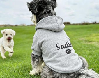 Może przedstawiać: Szara bluza z kapturem dla psa z imieniem "Sadie" i nadrukiem łapy. Pies siedzi na zielonej trawie, w tle biegnie inny pies. Bluza ma kaptur i prążkowane mankiety.