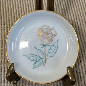 May include: A small, white ceramic dish with a gold rim and a gold and teal rose design. The dish is displayed on a brass stand. The rose design features a stylized flower and stem.