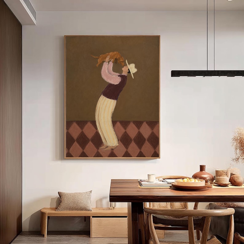 May include: A framed wall art print featuring a stylized illustration of a figure wearing a wide-brimmed hat, a dark vest over a pink long-sleeved shirt, and light-colored trousers, holding a small brown dog high above their head.