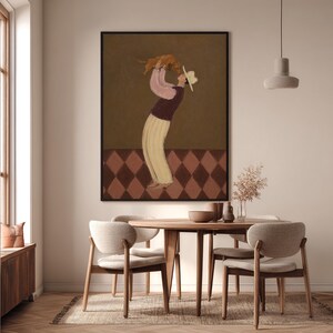 May include: A framed wall art print featuring a stylized illustration of a figure wearing a hat, a pink long-sleeved top, a dark vest, and cream-colored trousers, holding a small brown dog overhead against a dark brown background with a diamond-patterned floor.