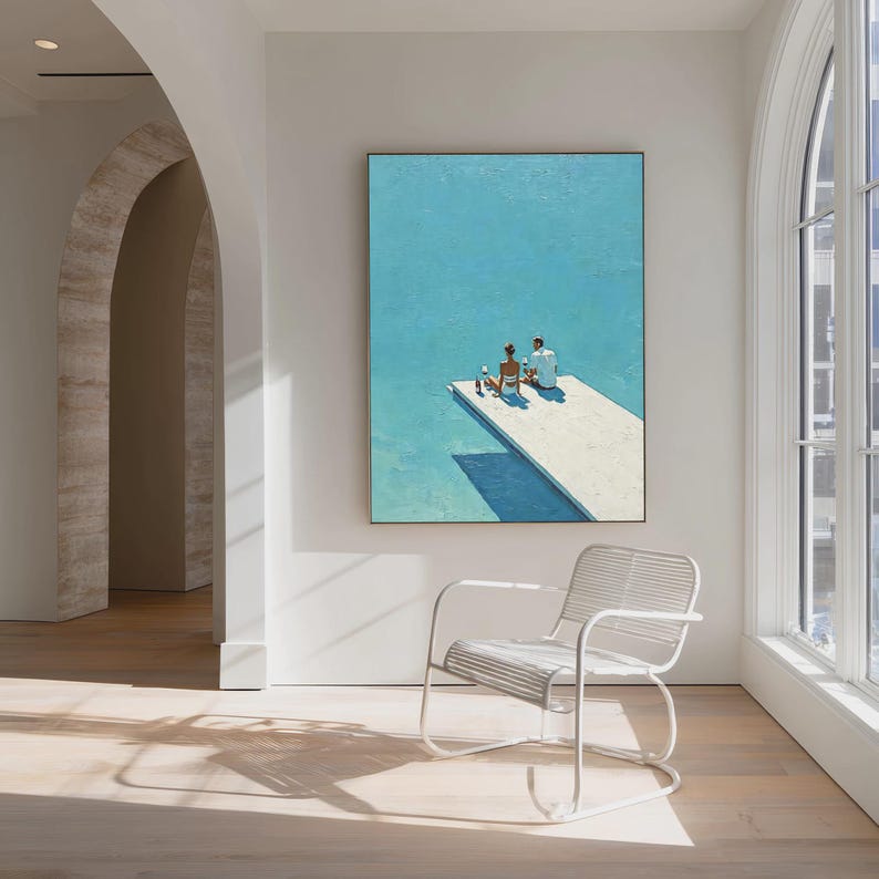 May include: A framed painting depicts two figures on a white diving board over a turquoise pool. The artwork is displayed on a white wall in a room with a white chair and wooden floor. Sunlight streams in from a large window.