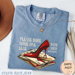 May include: Blue jean Comfort Colors t-shirt with the text "PLEASE BORE SOME ONE ELSE WITH YOUR QUESTIONS." A red high heel is printed on top of a stack of magazines. A gold necklace is draped across the shirt.